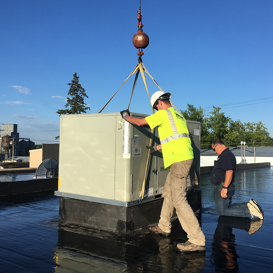 Technicians installing a large rooftop HVAC unit using a crane, with one worker guiding the equipment while another kneels nearby.