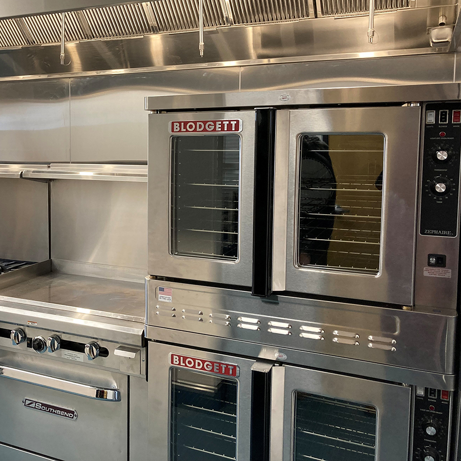 Commercial kitchen setup with stainless steel gas ranges, flat-top griddle, double Blodgett convection ovens, and overhead CaptiveAire ventilation hood.