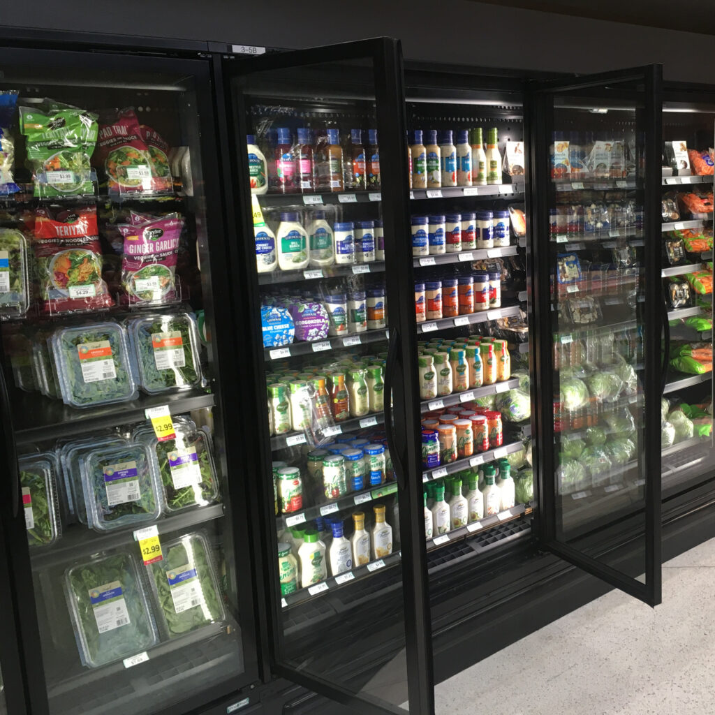 Supermarket refrigerated display cases filled with packaged salads, dressings, vegetables, and other fresh produce items.